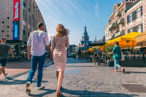 Mensen wandelen door een levendige straat met winkels en terrassen op een zonnige dag.