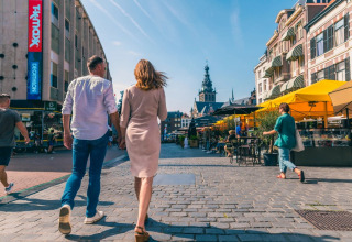 Folk går i en livlig by med butikker og caféer under solrige himmelstrøg i et ferieområde.