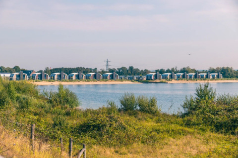Photo of glamping cabins by a lake in a holiday park, with lush greenery and a clear blue sky behind.
