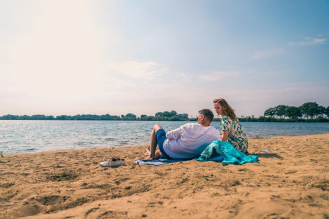 A couple relaxes on a sandy lakeside beach under a bright sky at a holiday park offering glamping stays.