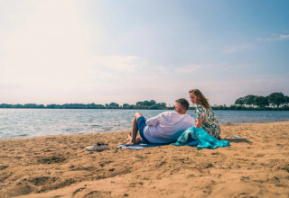 Et par slapper af på stranden ved en sø under en solrig himmel i en feriepark med glampingmuligheder.