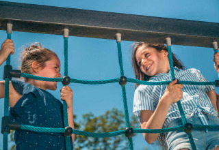 Twee kinderen klimmen samen lachend op een touwen klimrek tijdens een zonnige dag op een vakantiepark.