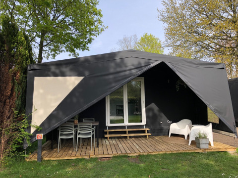 Tente safari noire nommée Coco Deluxe avec terrasse couverte, meubles de jardin et arbres verdoyants autour.