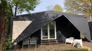 Schwarzes Safari-Zelt namens Coco Deluxe mit überdachter Terrasse, Gartenmöbeln und umgebenden Bäumen.