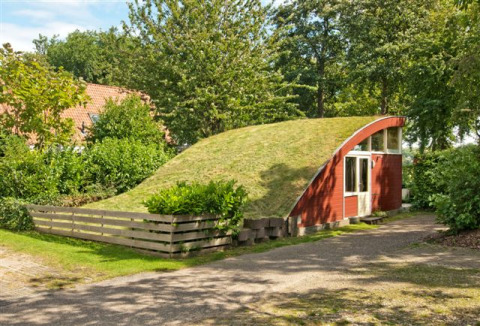 Kleine Grasdakbungalow met rond grasdak en rode gevels, omgeven door groene bomen en struiken.