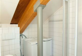 Lille badeværelse i Grasdakbungalow med brusekabine, vaskemaskine og synlige bjælker under skrå loft.