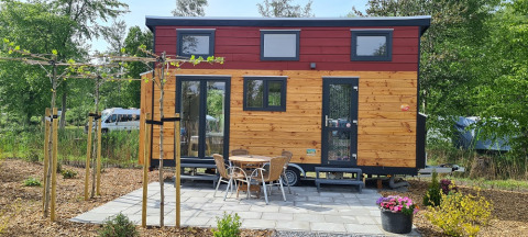 Pequeña casa de madera sobre ruedas con terraza, mesa y sillas, rodeada de árboles y plantas.