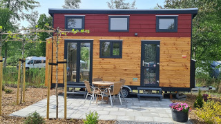Piccola casa in legno su ruote con patio, tavolo e sedie, circondata da alberi e verde.