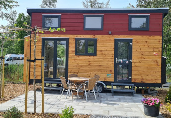 Pequeña casa de madera sobre ruedas con terraza, mesa y sillas, rodeada de árboles y plantas.