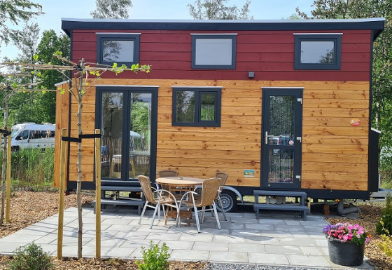 Tiny wooden house on wheels with patio, outdoor table and chairs, surrounded by trees and greenery.
