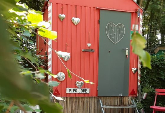 Kleine rote Tiny-House-Hütte namens Pipo Love, verziert mit silbernen Herzen, im Wald.