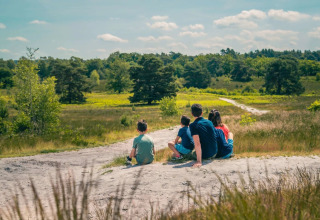 Een gezin zit op een zandige heuvel in een groen natuurgebied tijdens hun verblijf in een glamping vakantiepark.