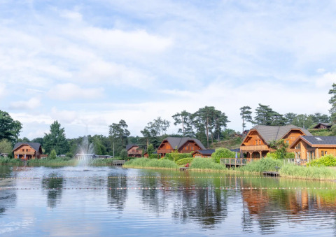 Luxe houten chalets aan een meer met fontein, omgeven door bomen in een vakantiepark voor glamping.