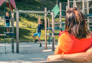 Een vrouw kijkt toe terwijl kinderen spelen op een klimrek in een vakantiepark met glamping accommodaties.