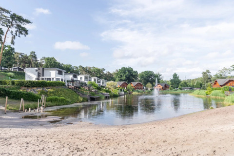 Zandstrand en meer met moderne en houten glamping accommodaties in een vakantiepark, omgeven door natuur.