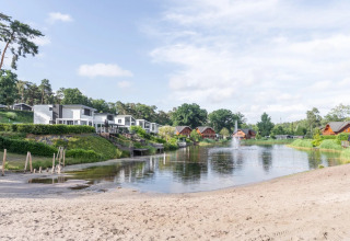 Zandstrand en meer met moderne en houten glamping accommodaties in een vakantiepark, omgeven door natuur.