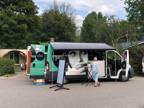 Foodtruck op een vakantiepark met een vrouw aan tafel, bomen en accommodaties op de achtergrond.