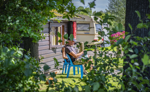 Un hombre se relaja en una silla azul frente a una caravana rodeada de árboles en Camping Batenstein, Utrecht.