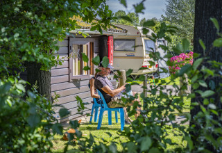 Een man zit op een blauwe stoel bij een caravan op Camping Batenstein in Utrecht, omringd door bomen.