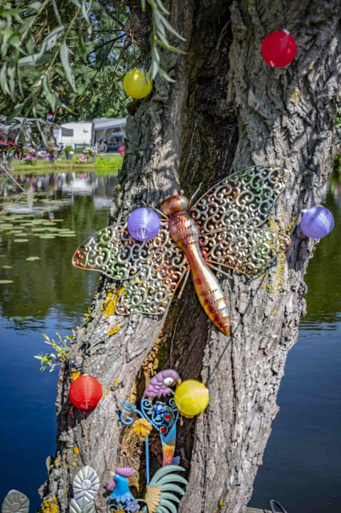 Árbol decorado junto a un estanque con una libélula metálica, farolillos y caravanas de fondo.