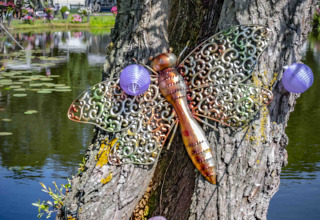 Versierde boom bij een vijver met metalen libel, kleurrijke lampionnen en caravans op de achtergrond.
