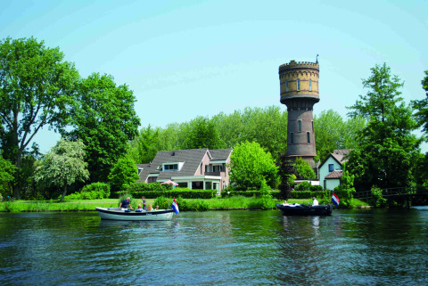 Barcos navegan por un río cerca de Woerden, Países Bajos, con una antigua torre de agua y casas rodeadas de árboles.