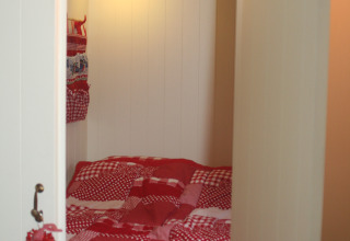 Acogedor rincón para dormir con edredón rojo en Pipowagen, Camping De Grienduil, Países Bajos.