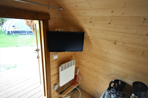 Interior of a tiny wooden house with a TV, heater, coffee set, and view of a tent outside the door.
