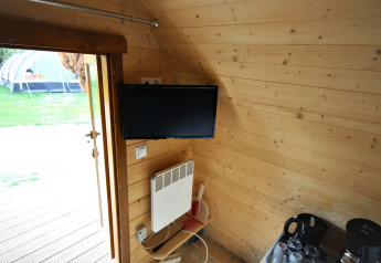 Interior of a tiny wooden house with a TV, heater, coffee set, and view of a tent outside the door.
