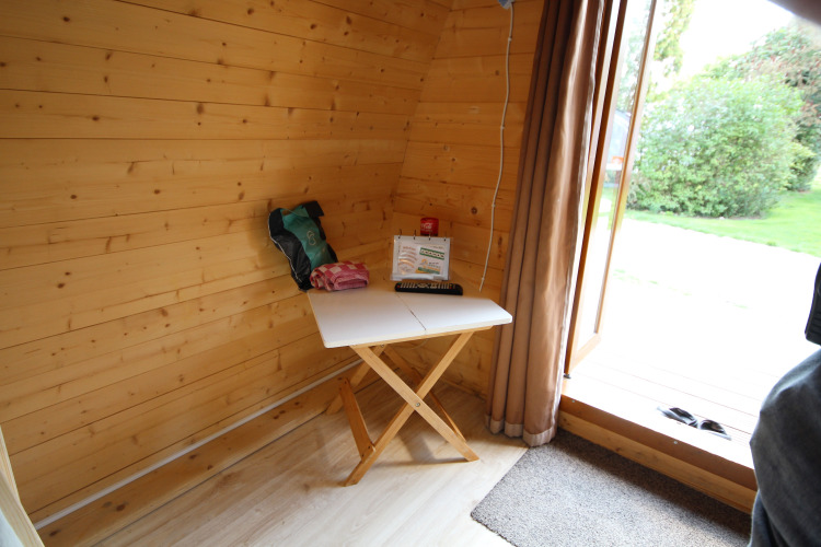 Interior de una pequeña casa de madera con una mesa, objetos encima y puerta abierta al jardín.