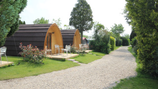 Twee houten Megapod VIP tiny houses met terrasstoelen bij een grindpad, omringd door groene beplanting.