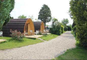 Twee houten Megapod VIP tiny houses met terrasstoelen bij een grindpad, omringd door groene beplanting.