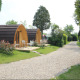 To charmerende trætinyhouses med små terrasser og havemøbler ved en grussti, omgivet af grønne planter.