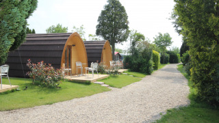 To charmerende trætinyhouses med små terrasser og havemøbler ved en grussti, omgivet af grønne planter.