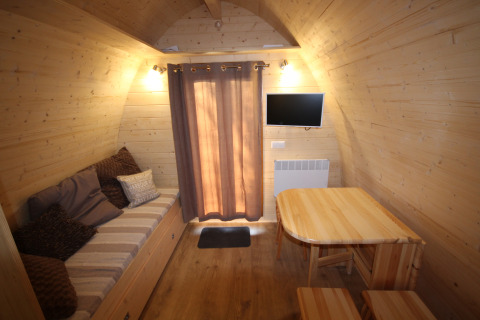 Interior de una casa pequeña de madera con sofá, mesa, taburetes, televisor y cortinas, Megapod VIP en Camping De Grienduil.