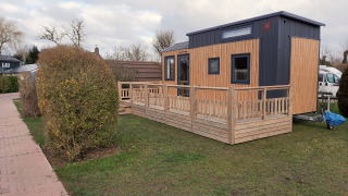 Ein kleines Holzhaus mit Veranda steht auf einer Wiese, umgeben von Büschen und Gehweg.