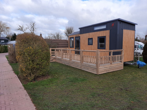 Een tiny house met houten terras op gras, aan een wandelpad en omgeven door struiken.