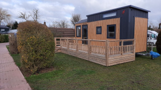 A tiny modern house with a wooden deck sits on a grassy yard next to a sidewalk and bushes.