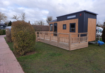 Una tiny house moderna con veranda in legno su prato, accanto a un sentiero e siepi.