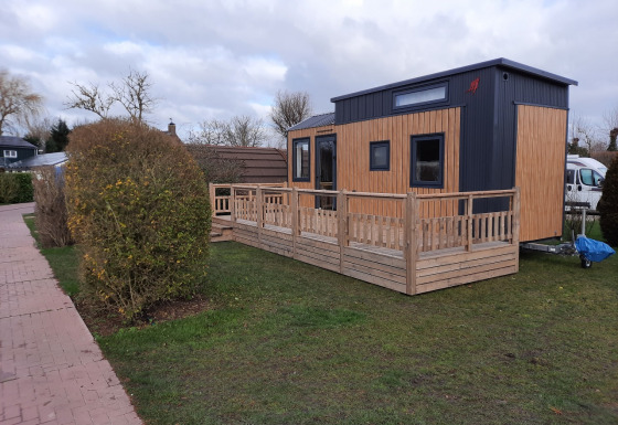 Una tiny house moderna con veranda in legno su prato, accanto a un sentiero e siepi.