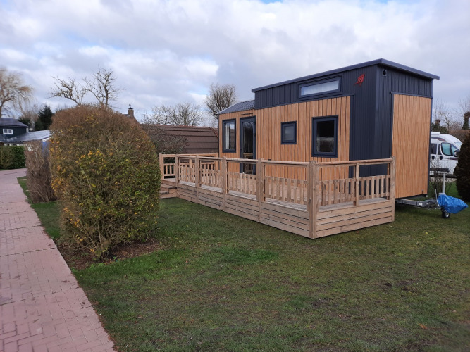 Een kleine moderne woning met houten terras op gras, naast een stoep en struiken.