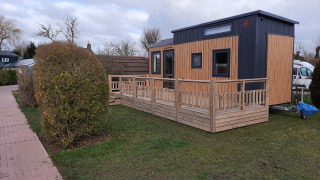 Una tiny house moderna con veranda in legno su prato, accanto a un sentiero e siepi.