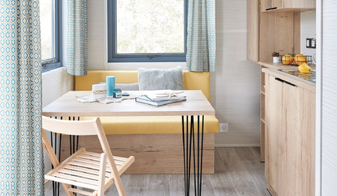 Interior luminoso y moderno de tiny house con mesa de madera, banco amarillo y ventanas en Camping De Grienduil.