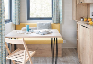 Interior luminoso y moderno de tiny house con mesa de madera, banco amarillo y ventanas en Camping De Grienduil.