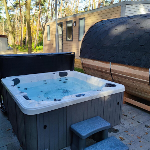 Jacuzzi exterior y sauna de barril frente a un lodge en Luxe Homes, Bospark De Brenkberg, Países Bajos, rodeados de bosque.