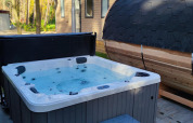 Jacuzzi exterior y sauna de barril frente a un lodge en Luxe Homes, Bospark De Brenkberg, Países Bajos, rodeados de bosque.