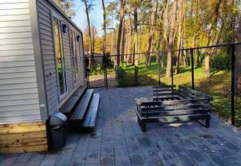 Modern lodge with an outdoor terrace and bench at Bospark De Brenkberg, Netherlands, surrounded by pine trees.