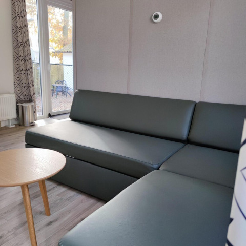 Moderne stue med grå sofa, rundt træbord og store vinduer i en lodge på Bospark De Brenkberg, Nederlandene.