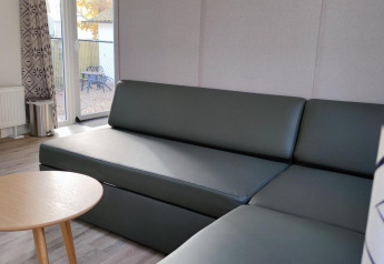 Modernes Wohnzimmer mit grauem Sofa, rundem Holztisch und großen Fenstern in einer Lodge bei Bospark De Brenkberg.