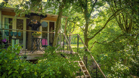 Treehouse surrounded by lush greenery at Buytenplaets Suydersee, Flevoland, Netherlands, with person on balcony.